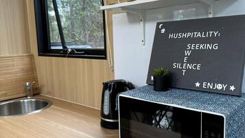 A kitchen with a microwave, a kettle, and a sink. There's a sign with the words 'HUSHPITALITY: SEEKING WISE SILENCE ENJOY'.
