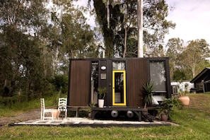 A small, dark-colored mobile home with a yellow door, situated on a gravel path with potted plants and a white chair nearby.