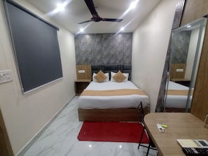 A hotel room with a bed, a desk, a chair, a mirror, and a ceiling fan. - Shree Sai Lodge (Prayagraj)