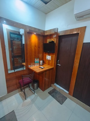 A compact kitchenette with a wooden countertop, a small chair, a trash bin, and a television mounted on the wall. - Shree Sai Lodge (Prayagraj)
