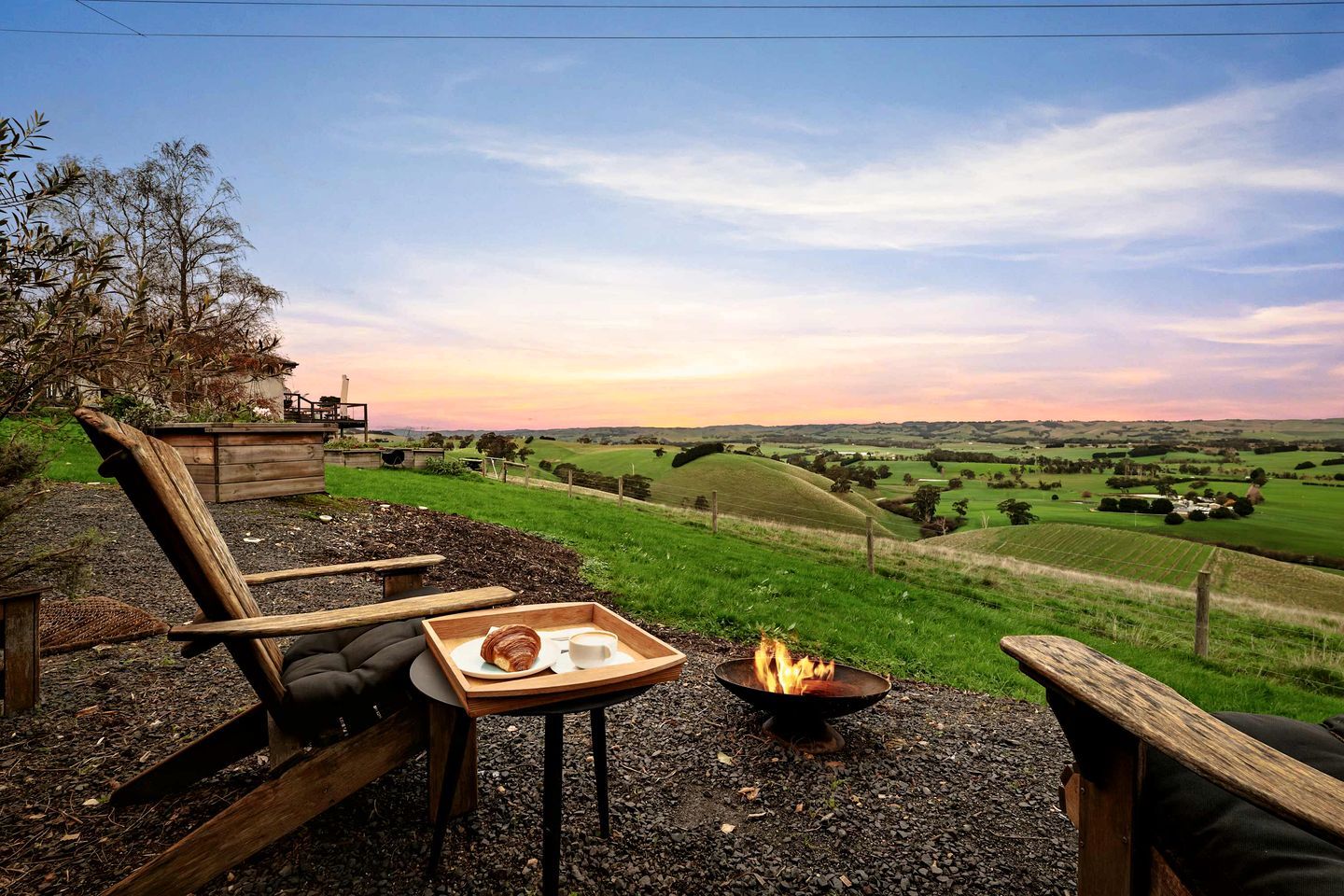 A rustic outdoor seating area with a fire pit, a tray with a croissant and a cup, and a view of rolling hills and fields.