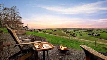 A rustic outdoor seating area with a fire pit, a tray with a croissant and a cup, and a view of rolling hills and fields.