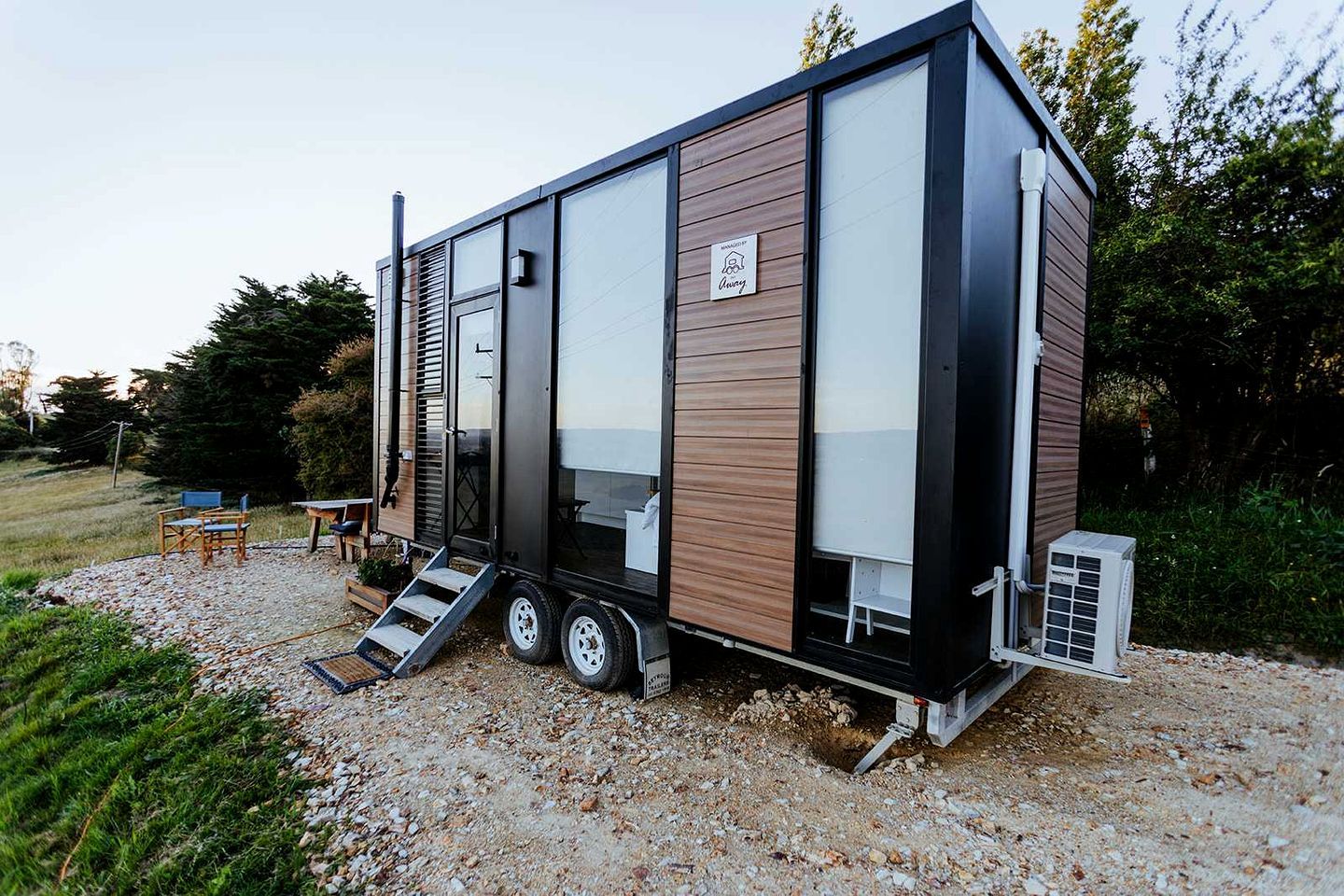 Une maison mobile moderne, dotée d’un toit plat, de grandes fenêtres et d’un petit espace extérieur aménagé pour s’asseoir.