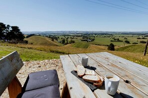 A wooden picnic table with two cups of coffee and slices of bread, overlooking a hilly landscape.