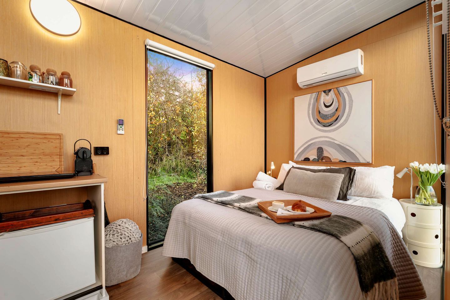 A modern bedroom with a bed, bedside table, and a view of greenery through a sliding door.