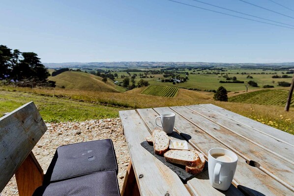 A wooden picnic table with two cups of coffee and slices of bread, overlooking a hilly landscape.