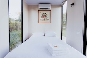 A hotel room with a bed, white linens, a framed artwork above the bed, and a view of the outdoors through large windows.