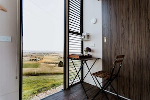 A room with a view of a rural landscape, a wooden table with a vase, and a chair.