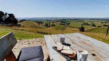 Ein Holztisch für ein Picknick mit zwei Kaffeetassen und Brotscheiben, mit Blick auf eine hügelige Landschaft.