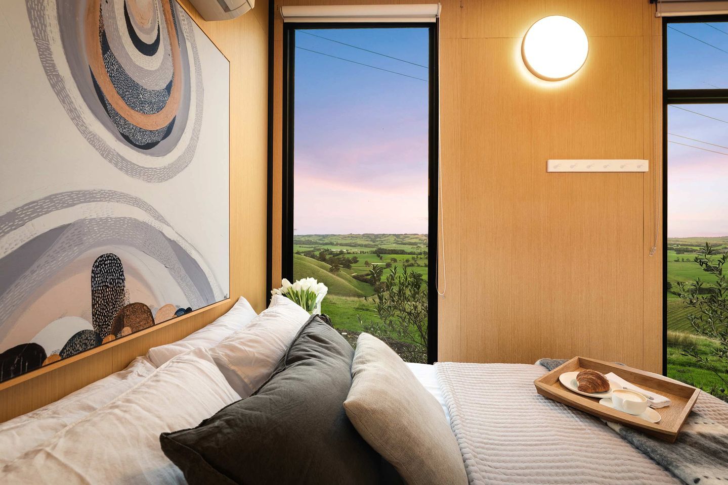 A bedroom with a large window, a bed with pillows, a bedside table with a tray of breakfast, and a wall with a circular light fixture.