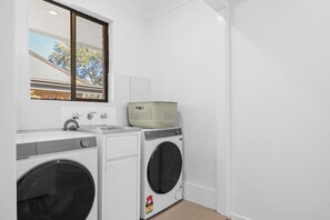 A laundry room with two front-loading washing machines, a sink, and a window with a view of a building and trees. - Hat Huts 40 Hutcheson St Hat Head (Hat Head)