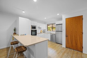 A modern kitchen with white cabinetry, a central island, bar stools, and a wooden door. - Hat Huts 40 Hutcheson St Hat Head (Hat Head)