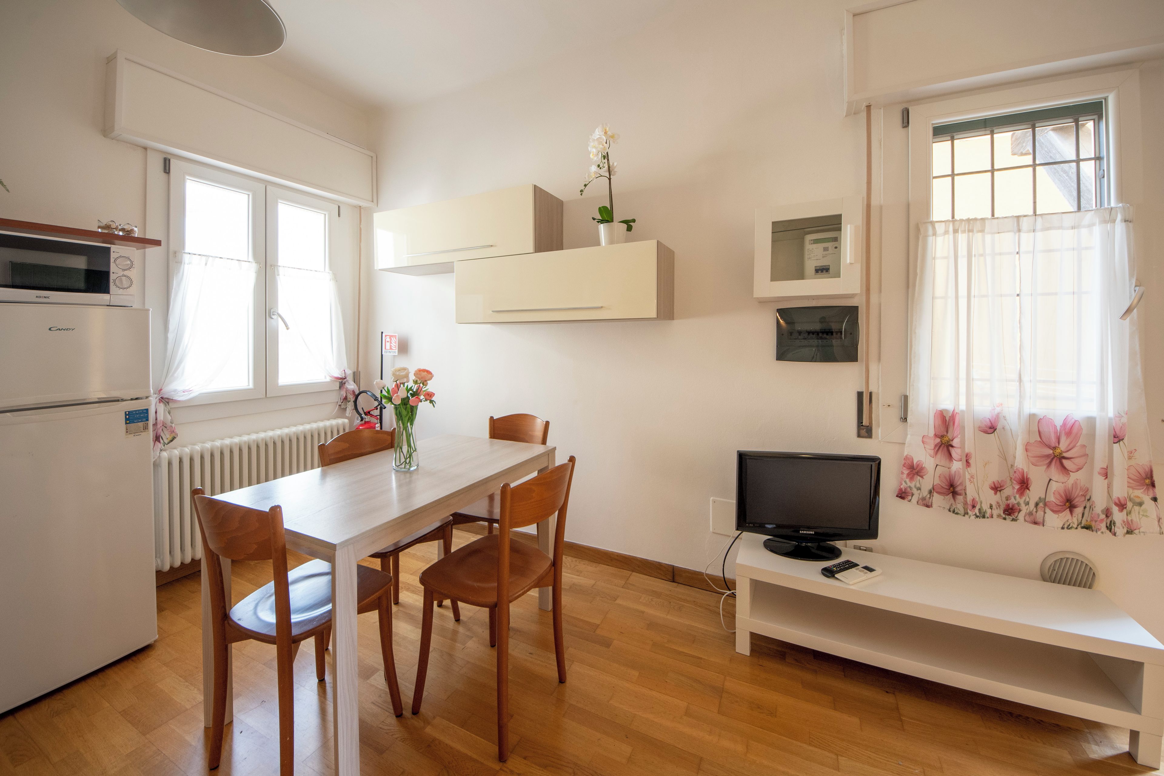 Una cocina moderna con electrodomésticos blancos, una mesa de comedor con sillas, un televisor sobre un mueble bajo y una ventana con cortinas estampadas.