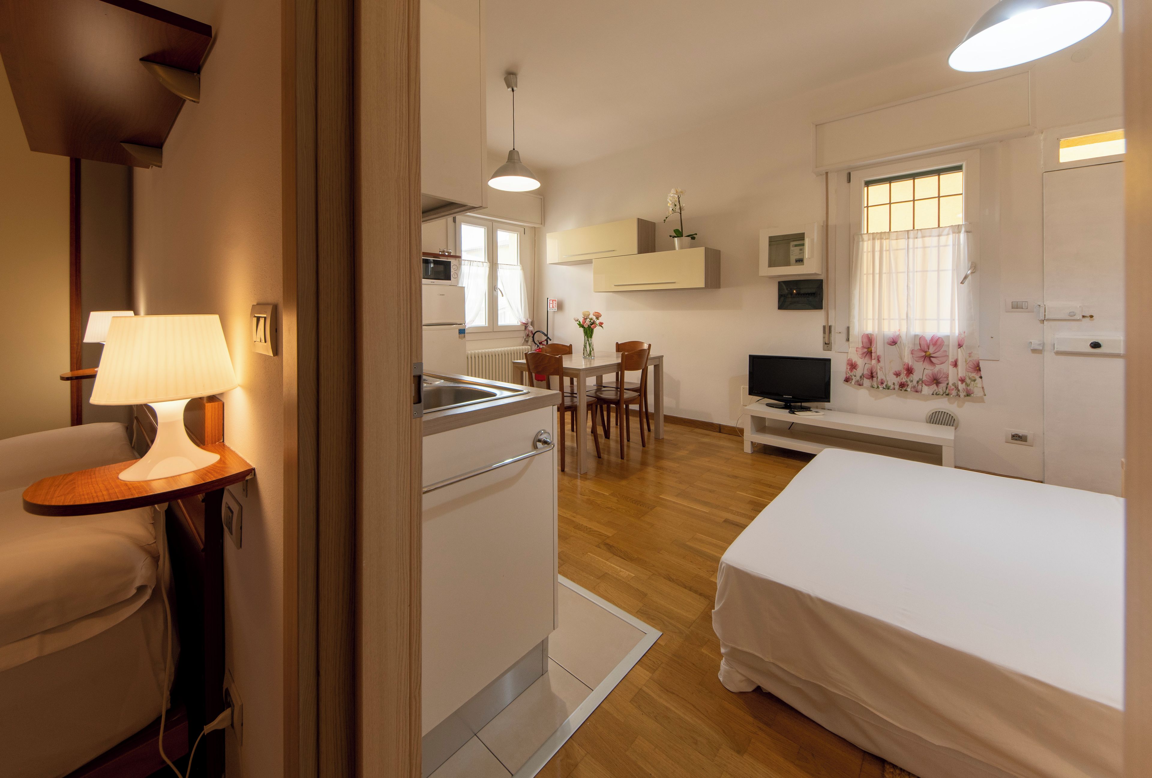 Habitación de hotel con cama, escritorio con lámpara, kitchenette, zona de comedor con sillas y televisión.