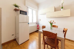 Cocina compacta con refrigerador, microondas y una pequeña planta en maceta. Hay una mesa de comedor con sillas y un florero con flores.