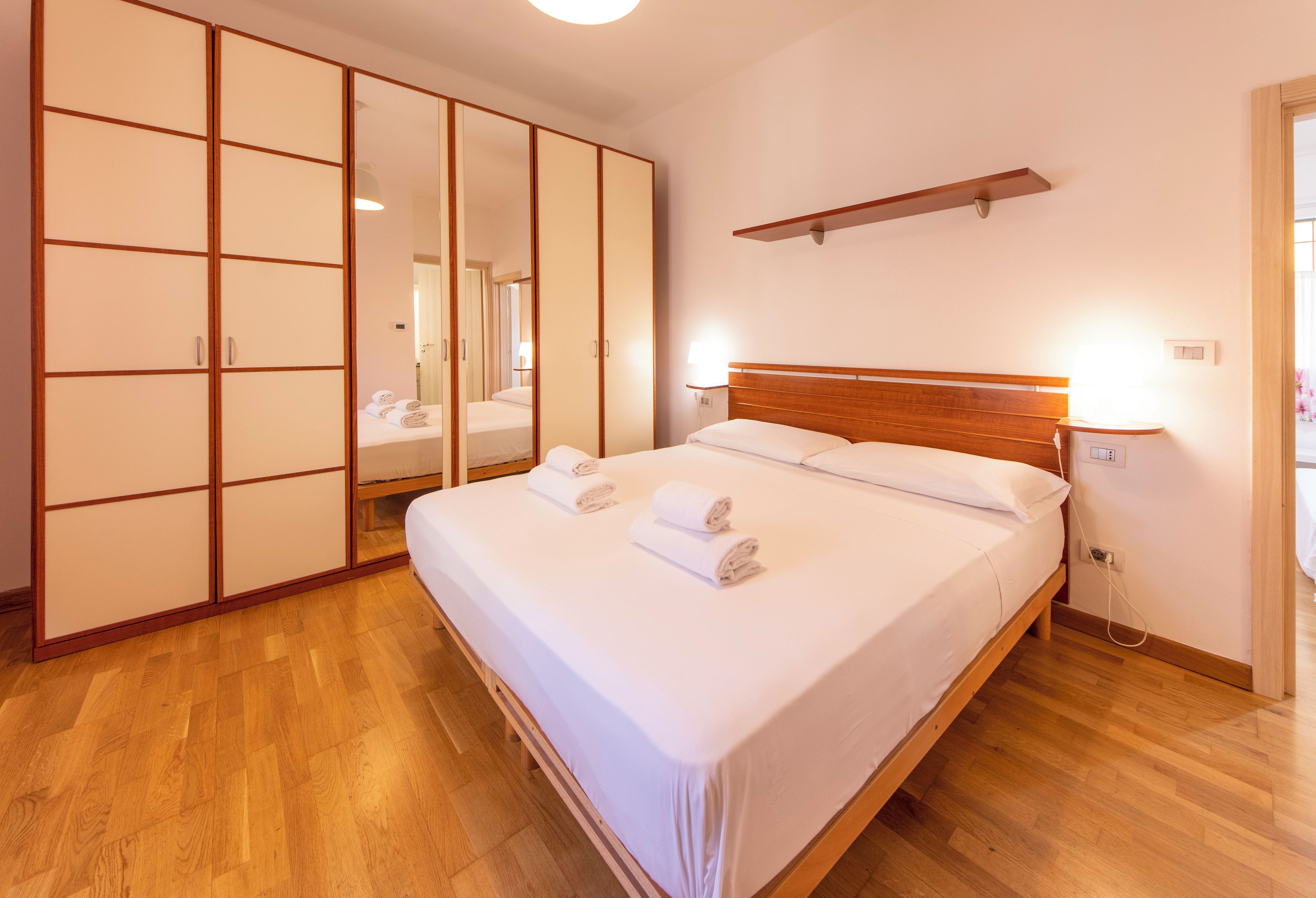 A hotel room with a bed, wooden flooring, a wardrobe with sliding doors, and a bedside table with a lamp.