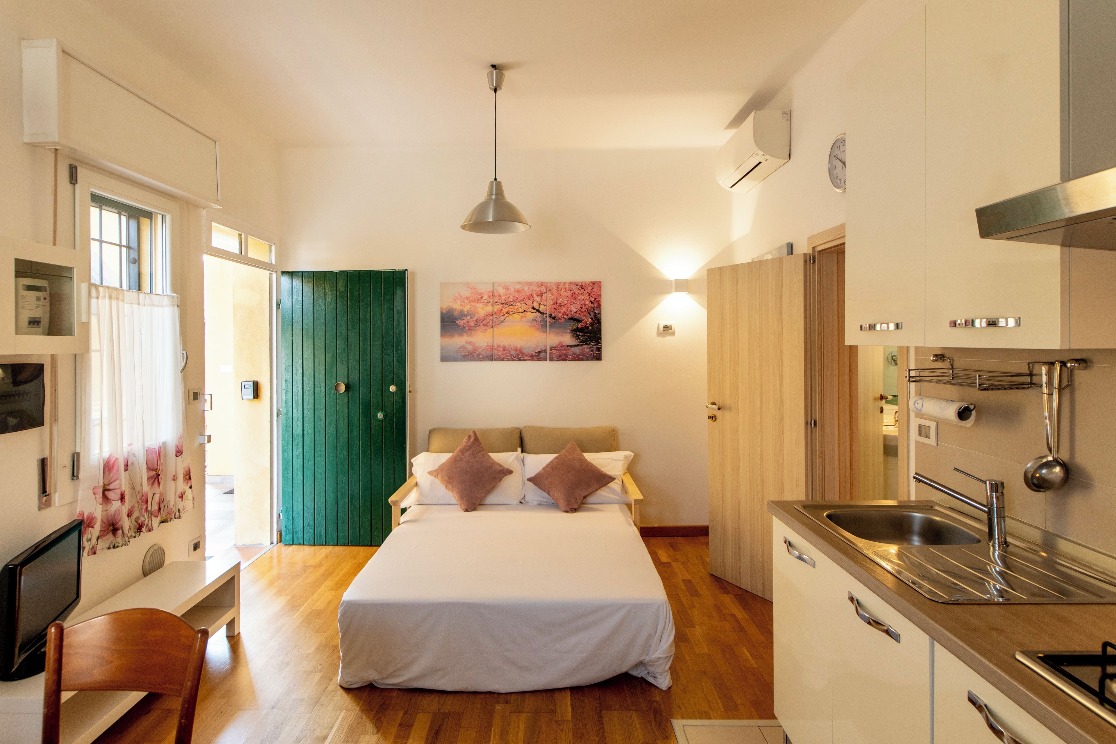 A bedroom with a bed, bedside tables, a green door, a painting, and a kitchen area with a sink.