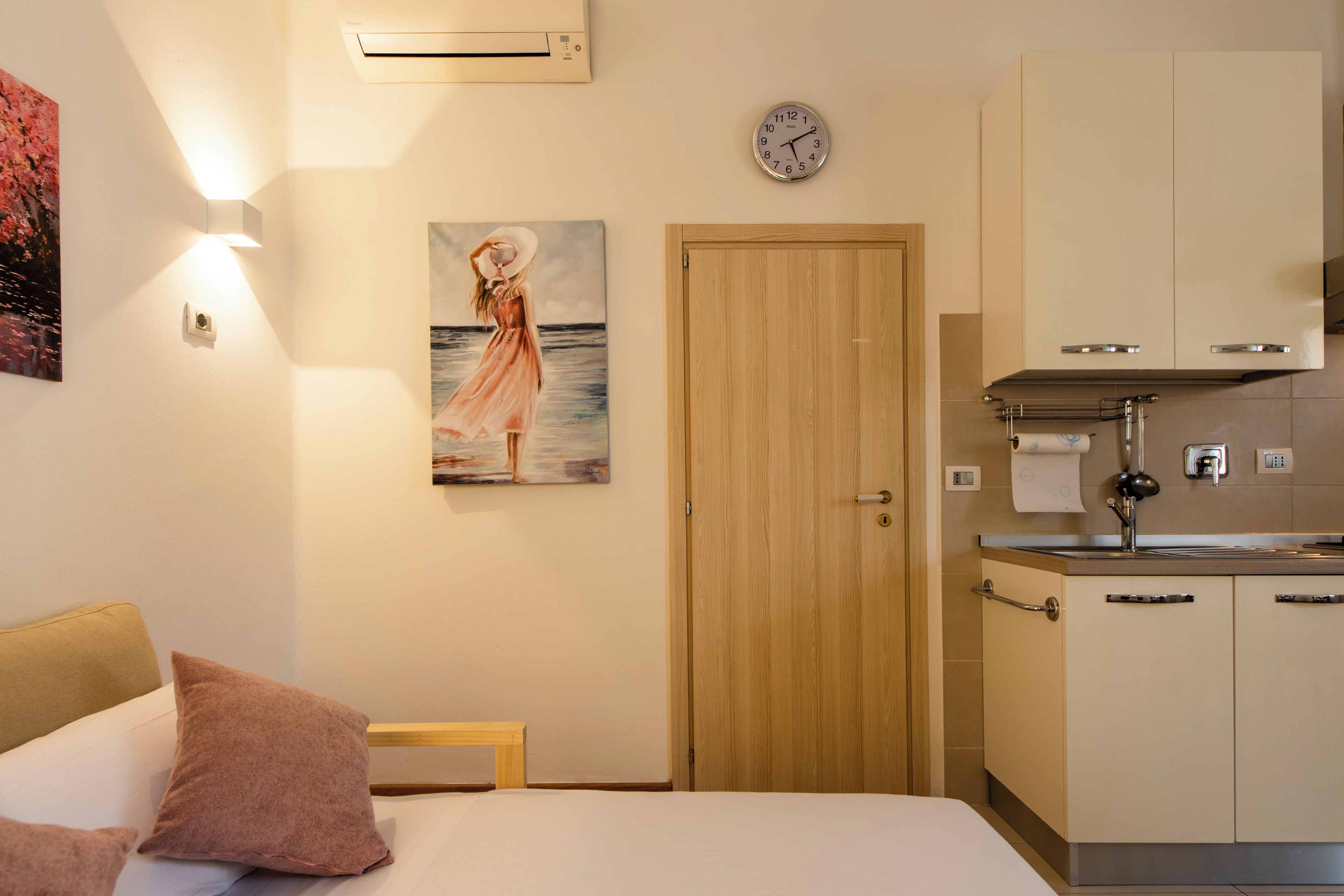 A hotel room with a bed, a painting, a clock, and a kitchenette.