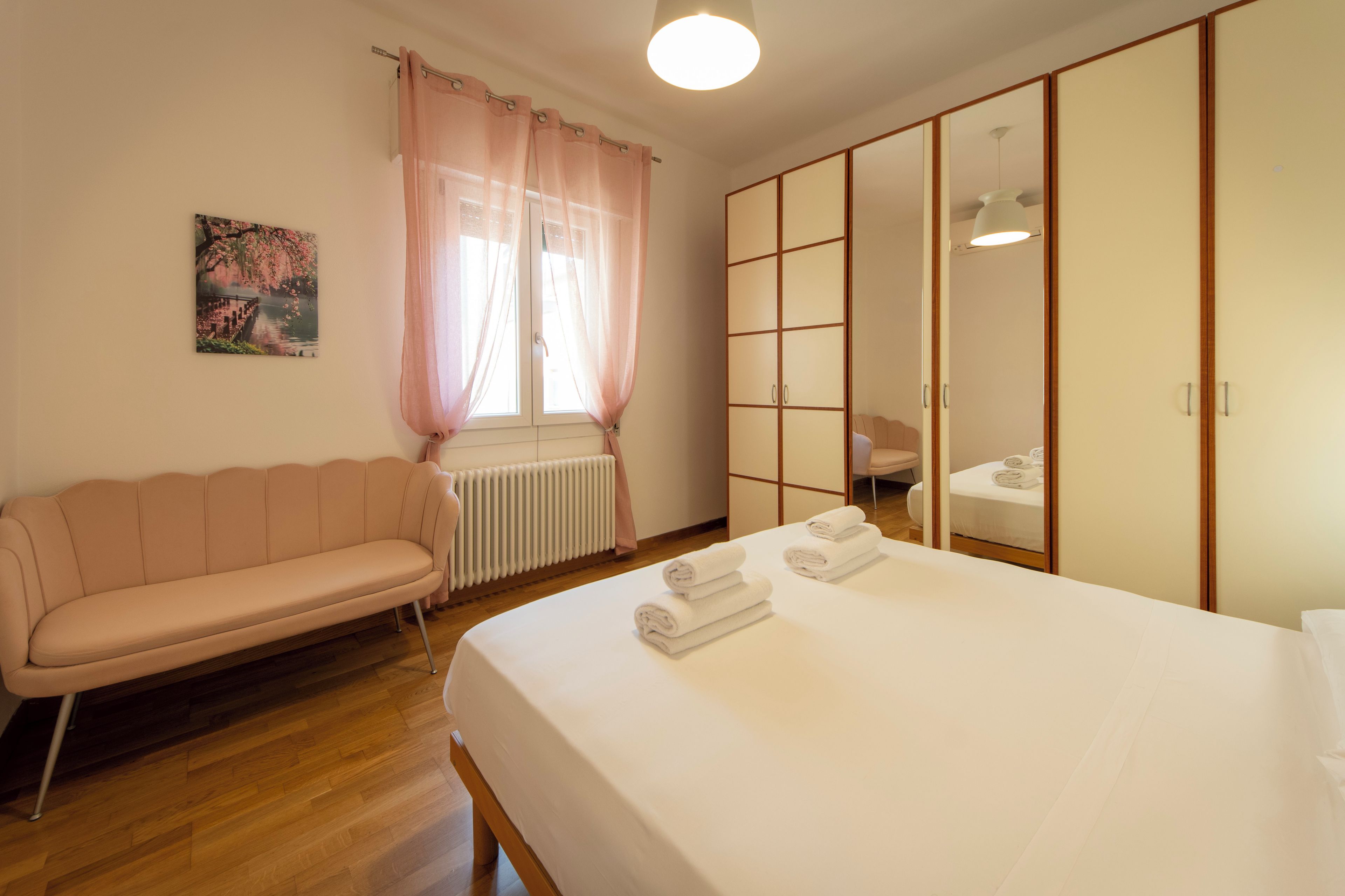 A hotel room with a bed, a sofa, a radiator, a window with curtains, a mirror, and a painting on the wall.