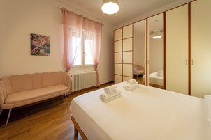 Habitación de hotel con cama, sofá, radiador, ventana con cortinas, espejo y un cuadro en la pared.