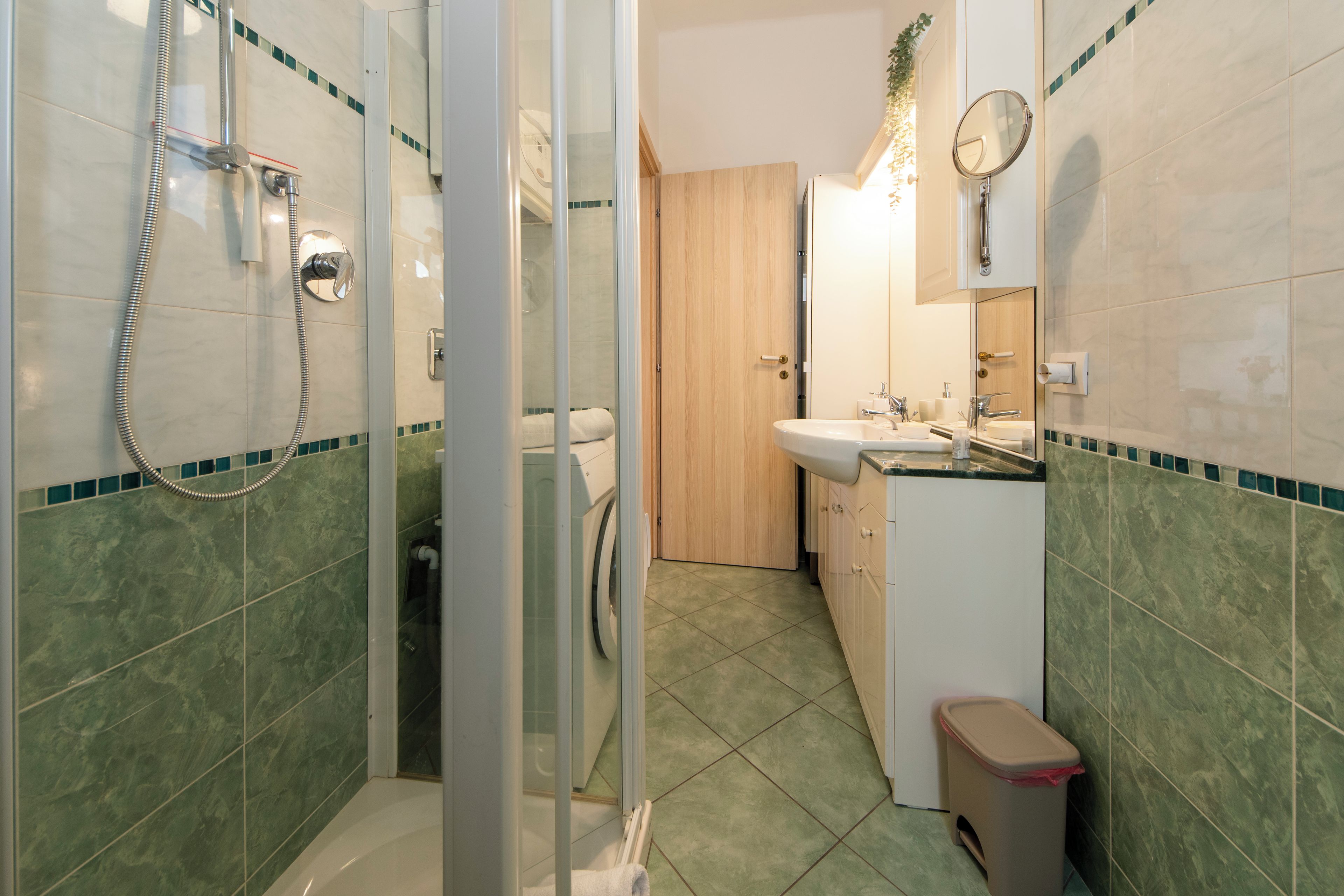 A bathroom with a glass shower enclosure, a toilet, a sink with a mirror, and a trash bin.