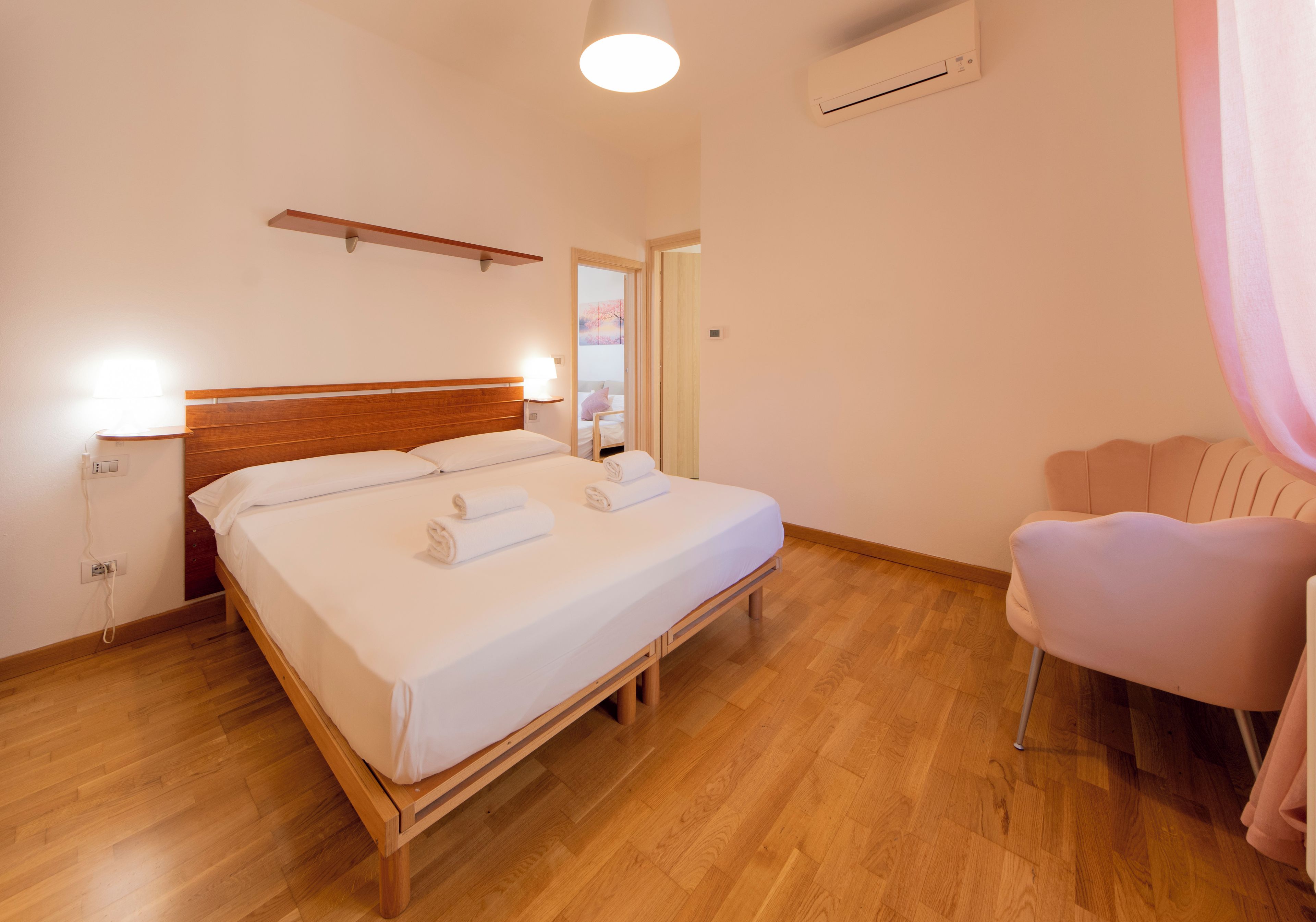 Habitación de hotel con cama de madera, lámparas de noche, una mesita, una silla y un equipo de aire acondicionado.