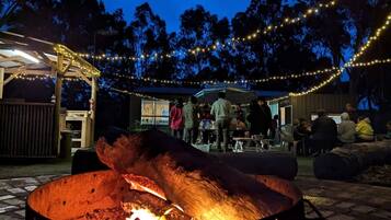 A fire pit with people around it, a covered seating area, and string lights in the background.