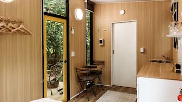 Quarto compacto com interior em madeira, uma pequena mesa com cadeiras, uma porta e janela com vista para o verde.