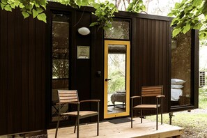 Una pequeña cabaña de madera con una puerta, dos sillas de madera y un banco en el exterior.