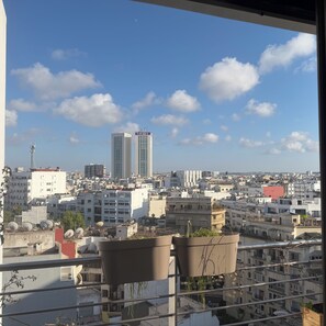 Eine Stadtansicht mit mehreren Gebäuden, darunter ein markanter Turm mit Beschilderung, aufgenommen von einem Balkon mit Blumentöpfen.