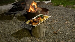 Foc de camp amb un plat de menjar i verdures a la brasa sobre un tronc de fusta.
