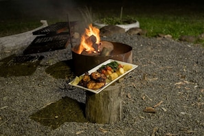 A campfire with a plate of grilled food and vegetables on a wooden stump.