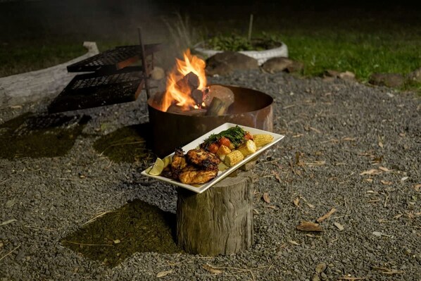 A campfire with a plate of grilled food and vegetables on a wooden stump.