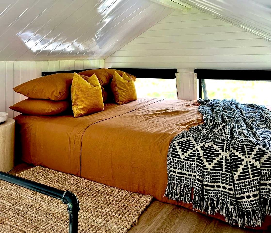 A bed with a brown bedspread and multiple pillows, a woven rug, and a patterned throw blanket.