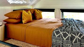 A bed with a brown bedspread and multiple pillows, a woven rug, and a patterned throw blanket.