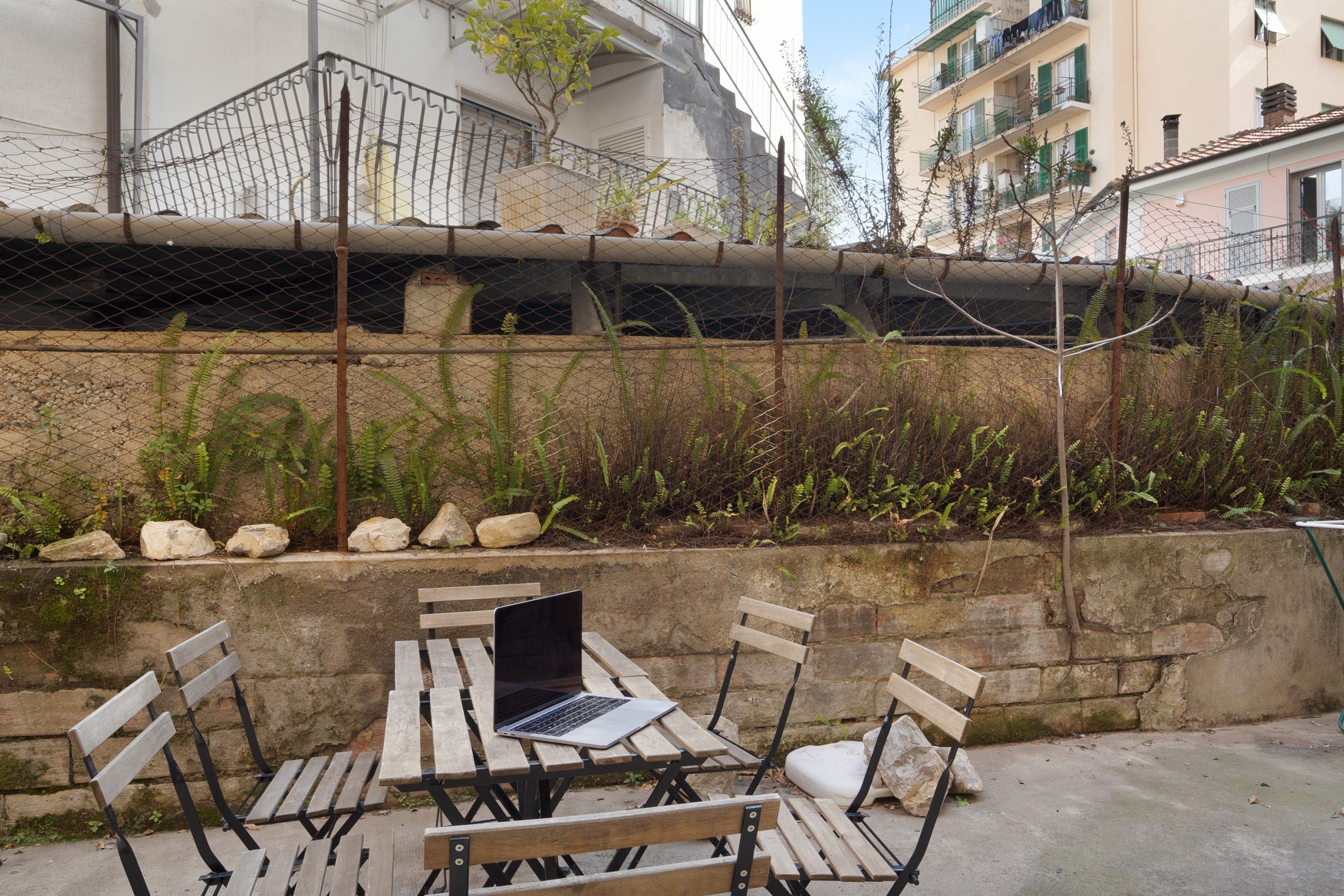 An outdoor seating area with a table and chairs, a laptop on the table, and a stone wall with plants and rocks.