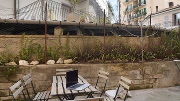 An outdoor seating area with a table and chairs, a laptop on the table, and a stone wall with plants and rocks.