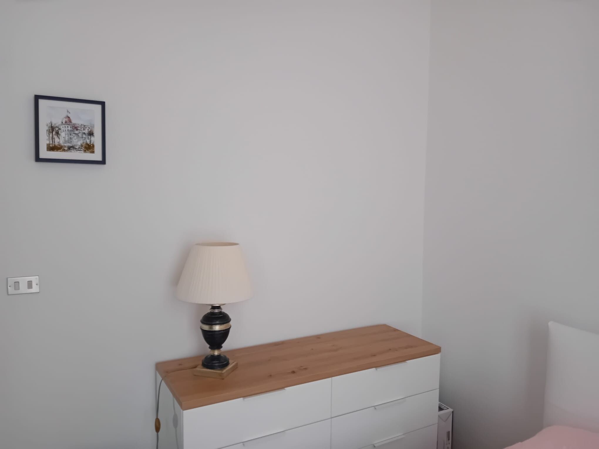 A bedroom with a wooden nightstand, a white dresser, a bedside table with a lamp, and a framed picture on the wall.
