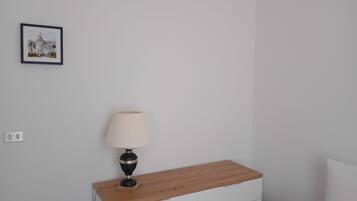A bedroom with a wooden nightstand, a white dresser, a bedside table with a lamp, and a framed picture on the wall.