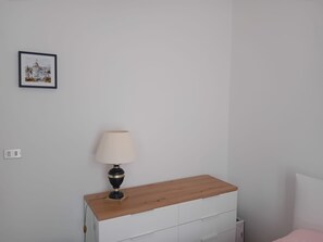 A bedroom with a wooden nightstand, a white dresser, a bedside table with a lamp, and a framed picture on the wall. - Il Gattino di Sanremo 1 – Relaxing Apartment with Wi-Fi, AC & Parking (Sanremo)