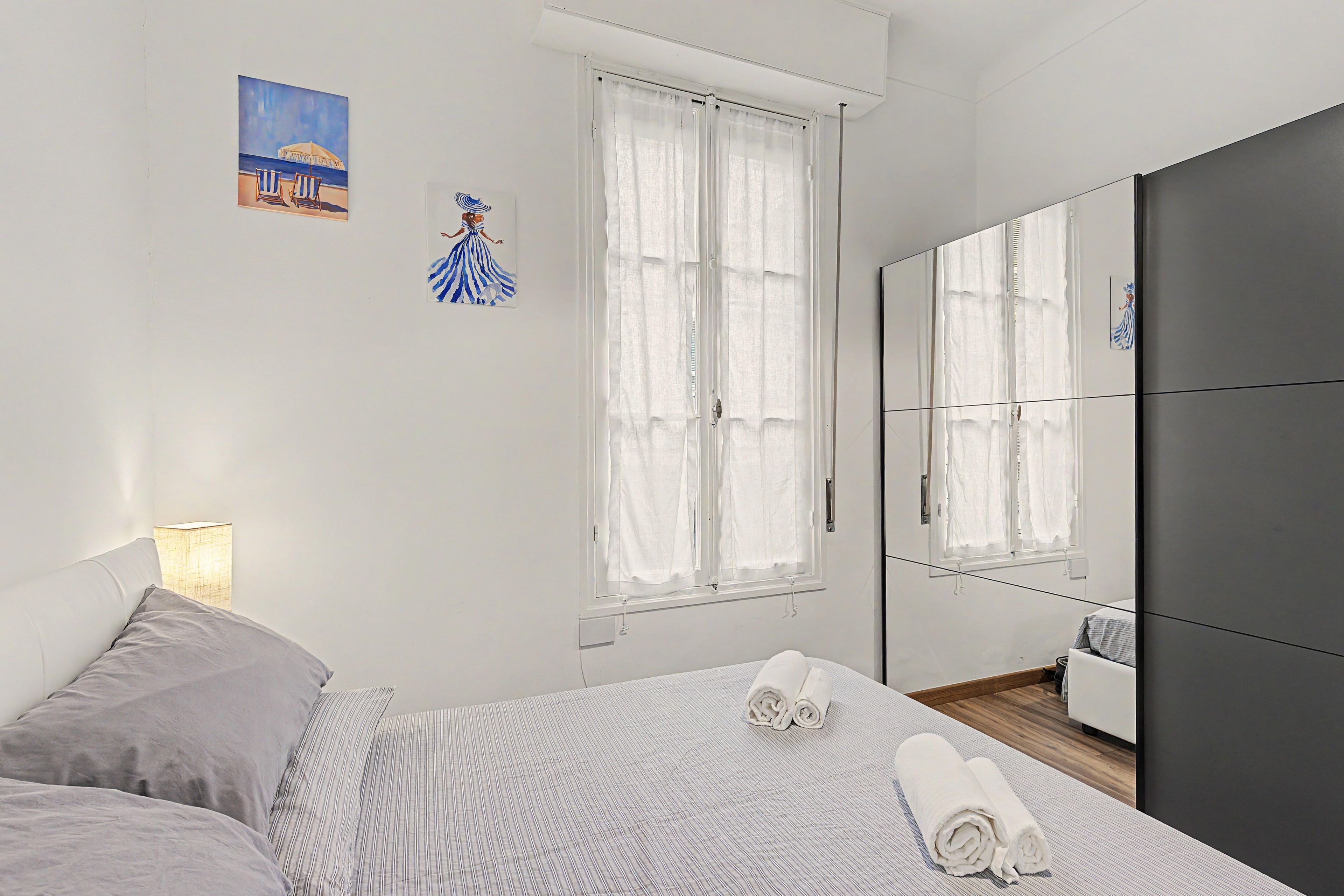 Une chambre à coucher avec un lit, un grand miroir, une fenêtre avec des rideaux et un tableau accroché au mur.