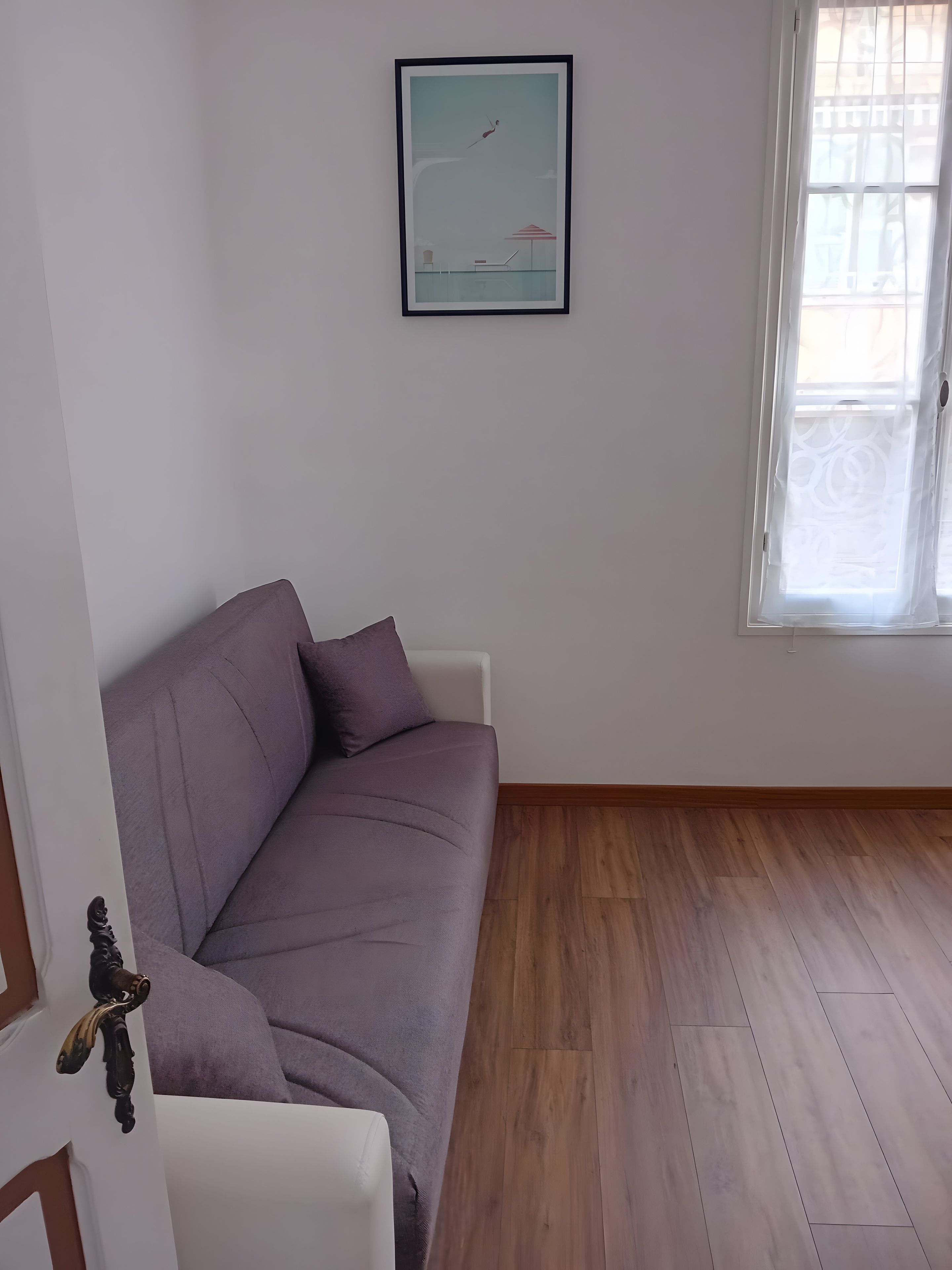 A room with a purple sofa, a framed picture on the wall, and a window with curtains.