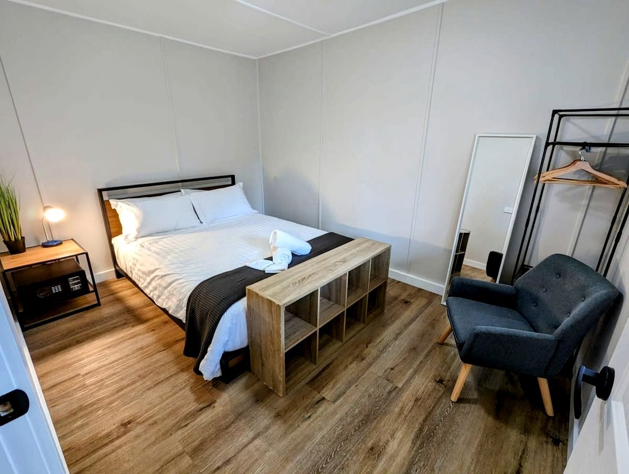 A bedroom with a bed, bedside table, bench, chair, and a hanging clothes rack.