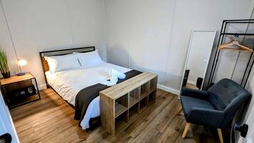 A bedroom with a bed, bedside table, bench, chair, and a hanging clothes rack.