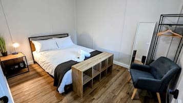A bedroom with a bed, bedside table, bench, chair, and a hanging clothes rack.