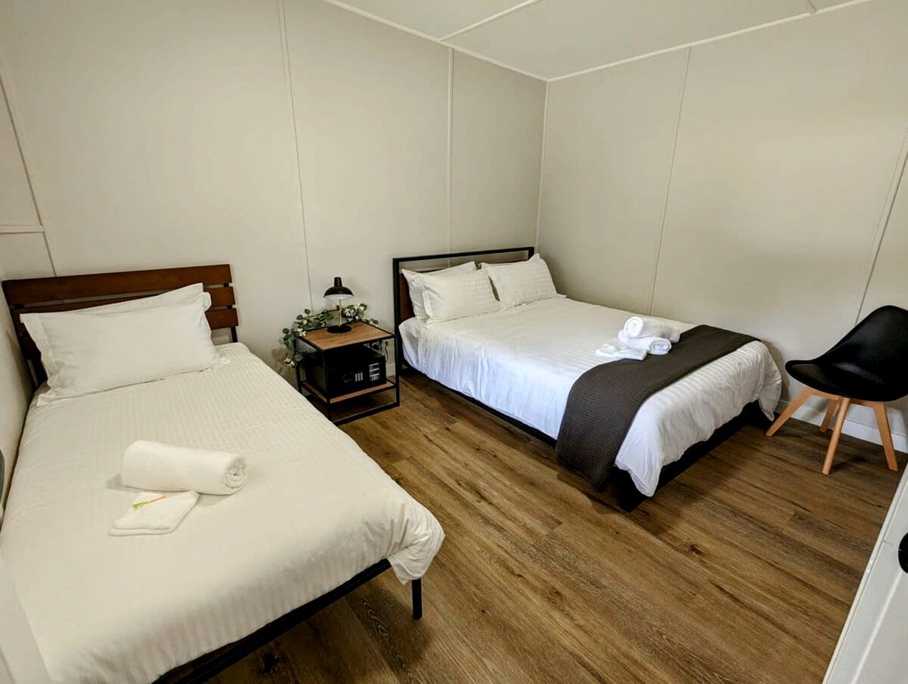 A hotel room with two beds, a bedside table with a lamp, a chair, and a small plant.