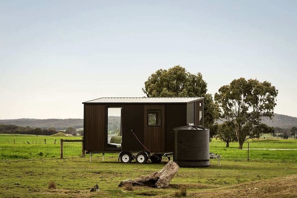 A small, dark-colored cabin with a sliding door, a water tank, and a trailer attached.