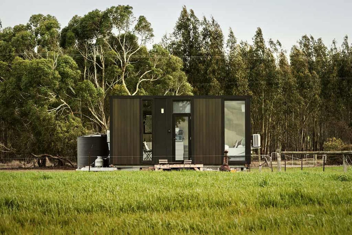 A modern, single-story cabin with a large sliding glass door, situated in a grassy field with trees in the background.