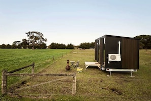Une petite maison mobile de couleur sombre, dotée d’un climatiseur blanc, est située dans un champ verdoyant, avec une clôture en bois en premier plan.