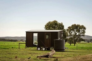 A small, dark-colored cabin with a sliding door, a water tank, and a trailer attached.